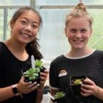 Two high school students smile for a photo while holding small plants in a greenhouse.